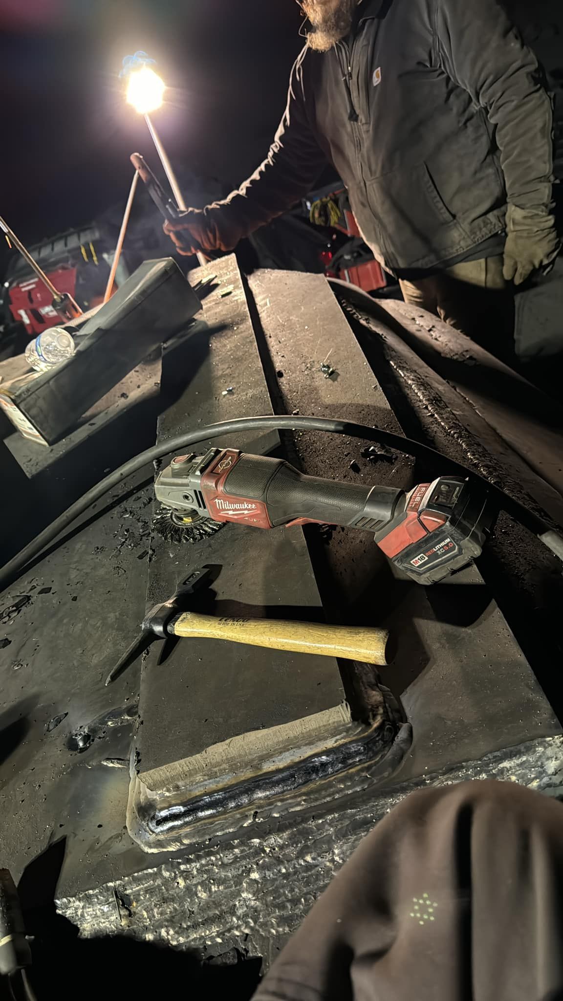 Man working on a dark surface at night with a hammer and power tool. Bright light overhead.