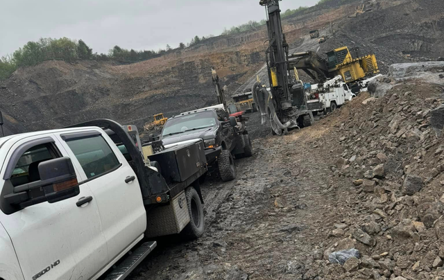 White truck towing heavy machinery on a rocky, industrial site, gray sky overhead.
