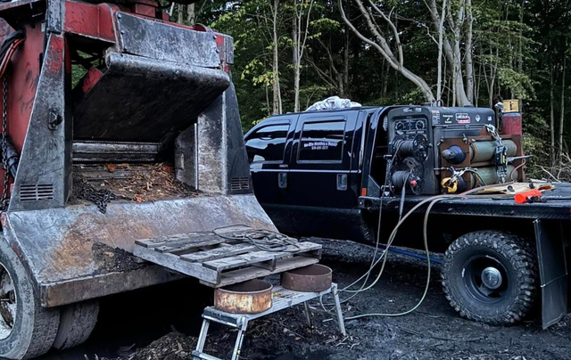 A wood chipper next to a truck with welding equipment in a wooded area.