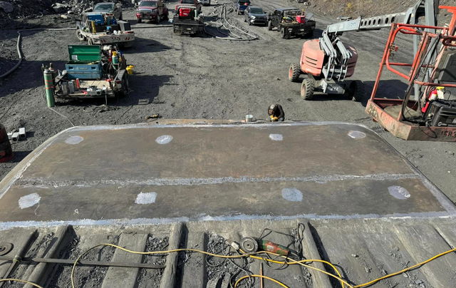 Overhead view of a large metal plate with welded patches at a construction site, vehicles in background.