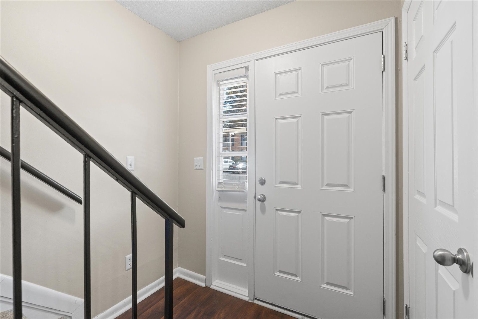 Interior view of a light-walled entryway with a white six-panel door, an adjacent side light, and a dark metal railing.
