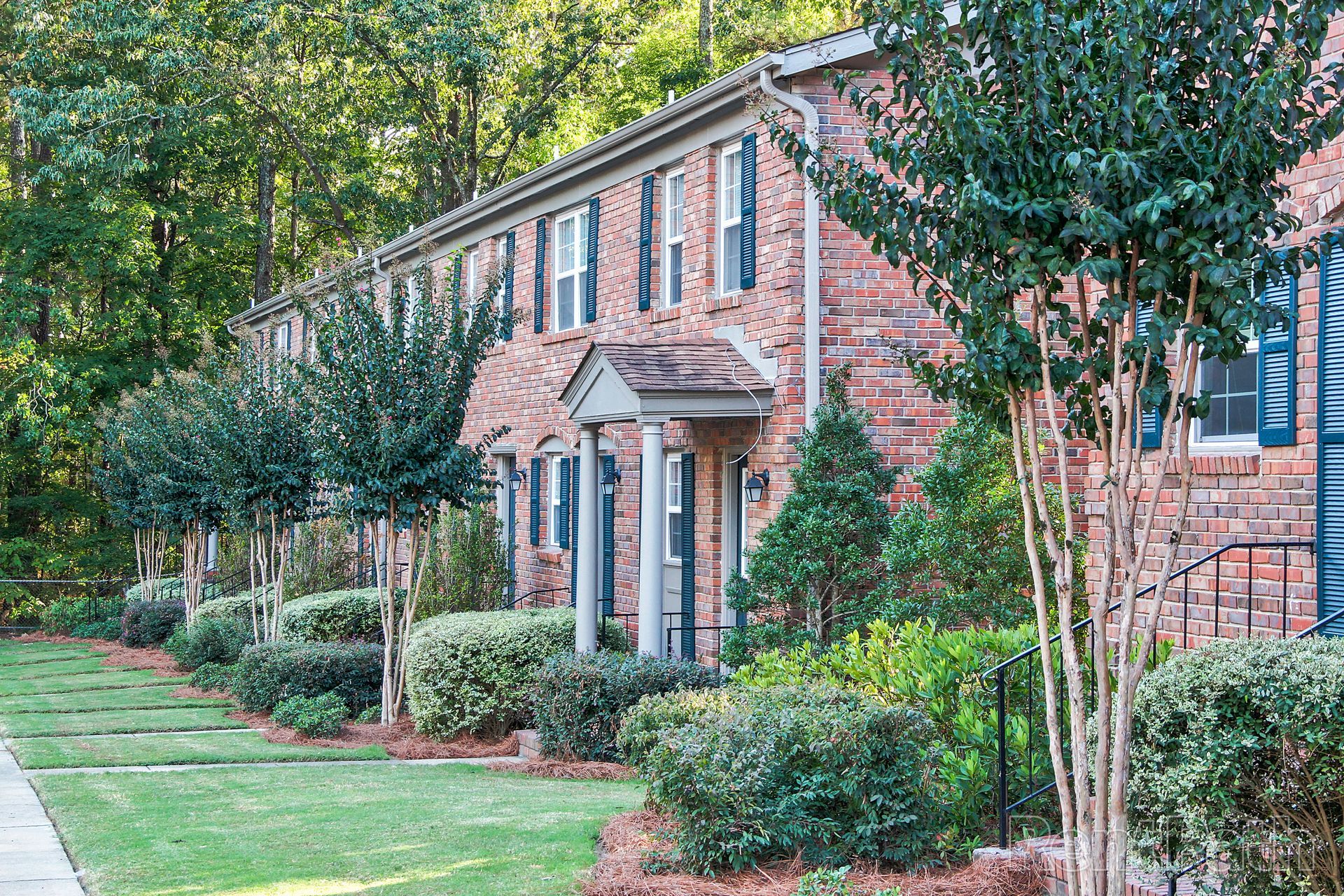 A multi-story brick townhouse building with front porches, surrounded by manicured shrubs and trees on a sunny day.
