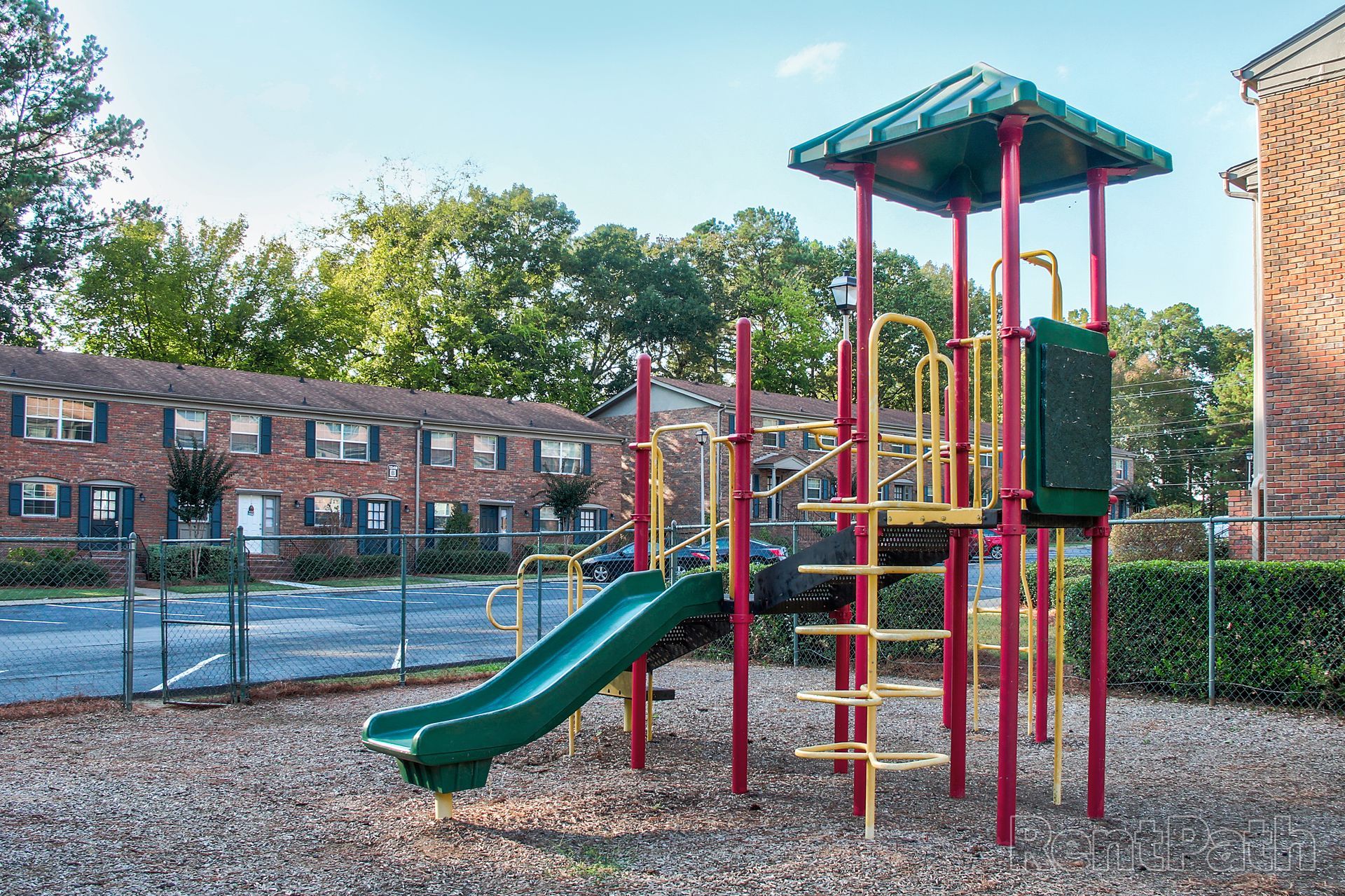 A colorful playground structure with a green slide, yellow ladders, and a green roof, set on wood chips near a brick building.
