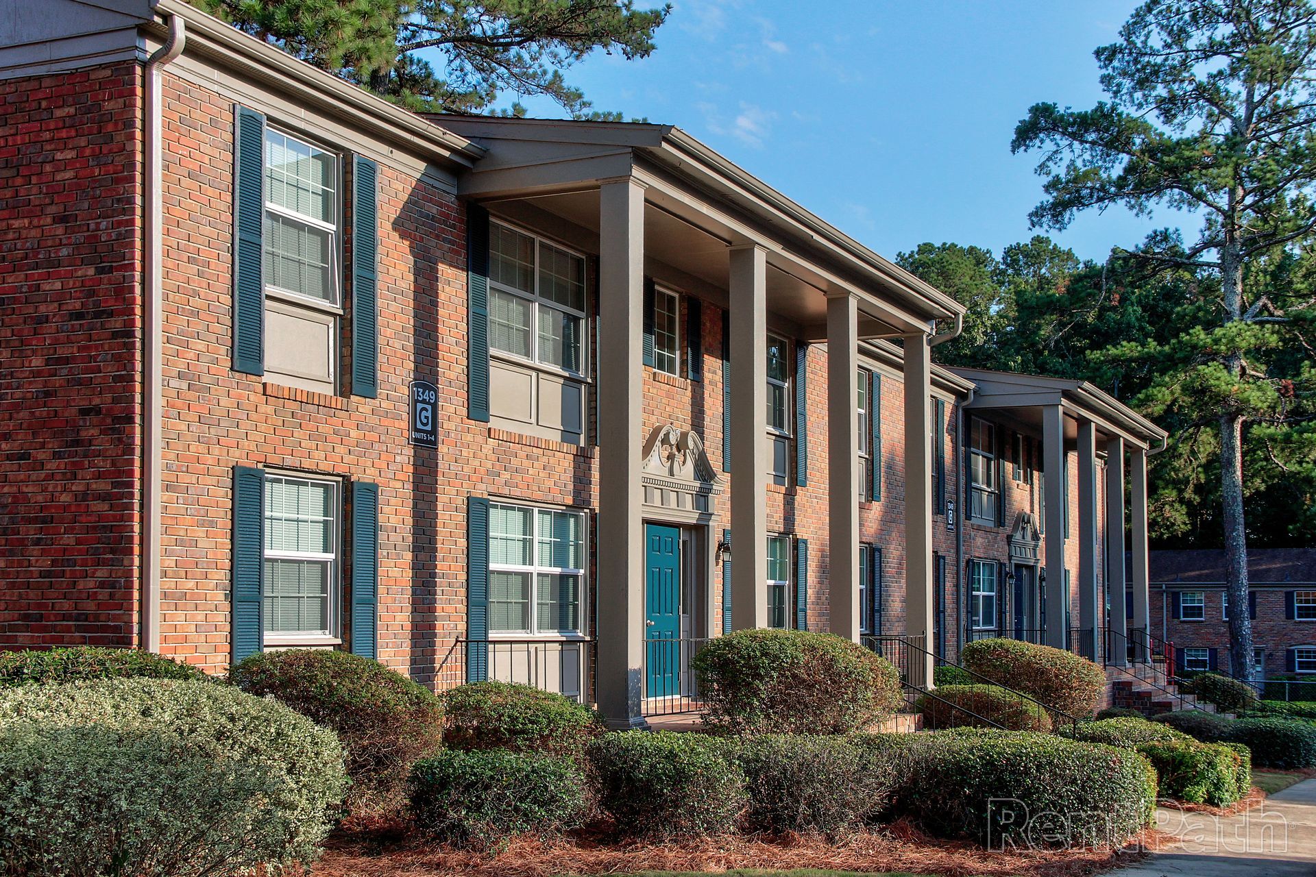 A two-story brick apartment building with tall, white columns, blue shutters, and rounded shrubbery in the foreground.