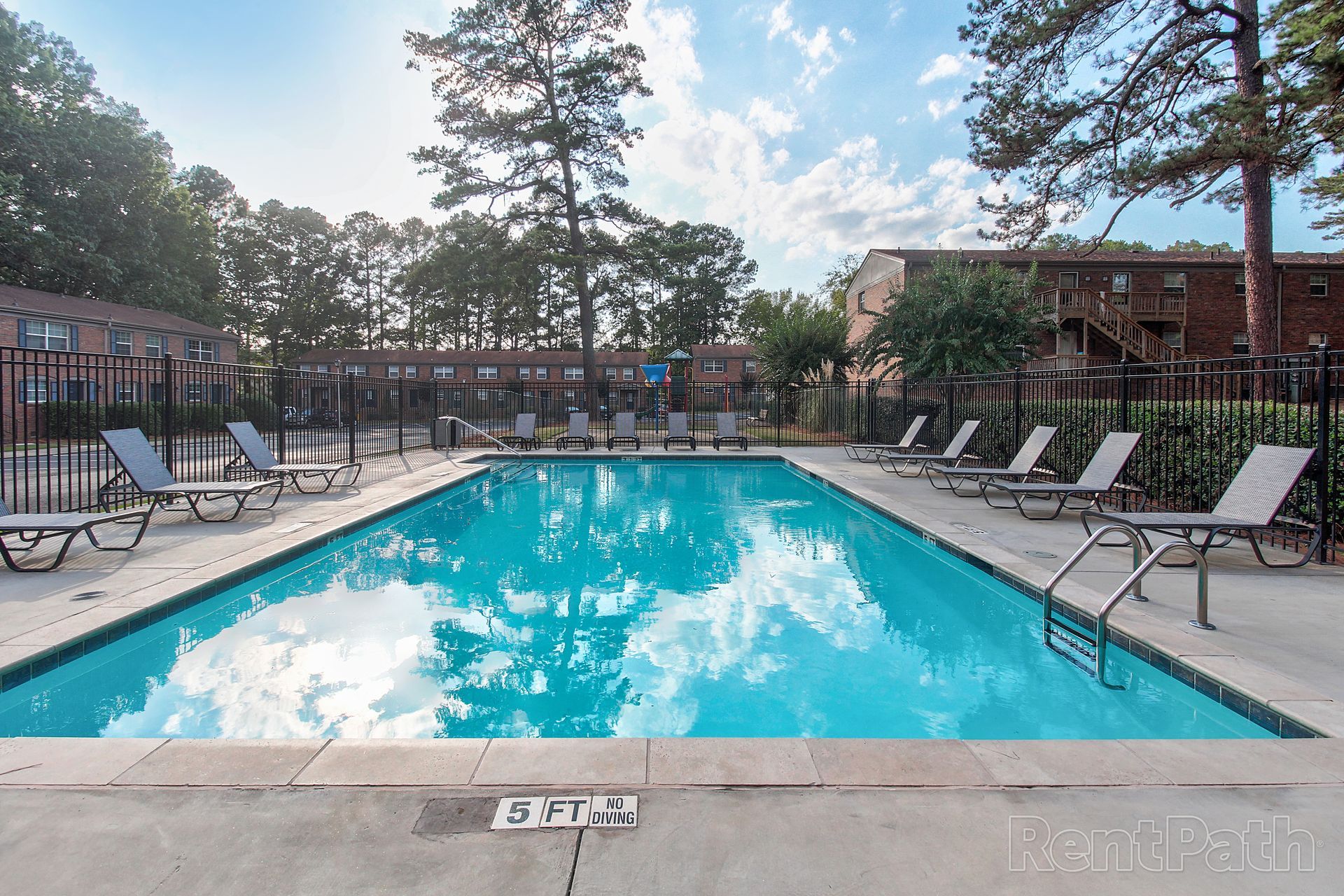 A rectangular outdoor swimming pool surrounded by a concrete deck with lounge chairs and trees in the background.