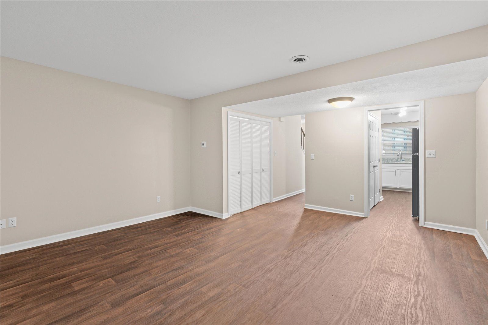 A bright, unfurnished room with light beige walls and wood-patterned flooring, leading toward a kitchen doorway.