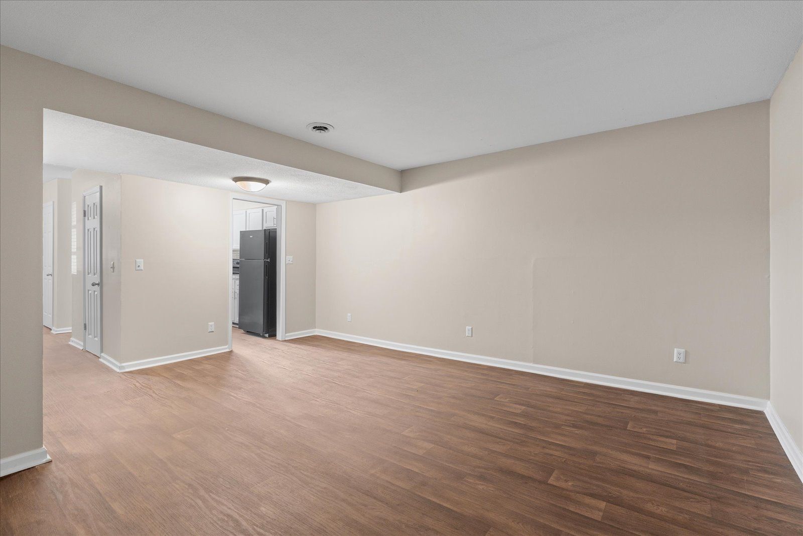 A brightly lit, empty room with wood-look flooring, light beige walls, and an open doorway leading to a kitchen area.