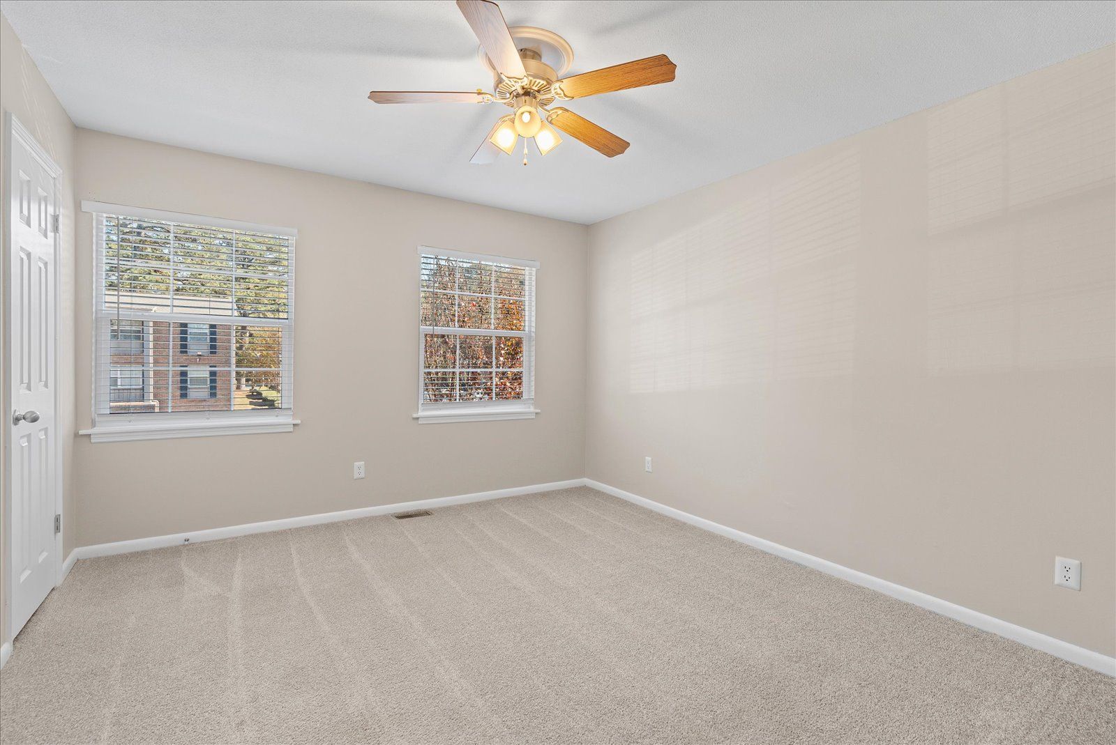 An empty bedroom with neutral beige walls, light carpet, a ceiling fan, and two windows looking out at trees.