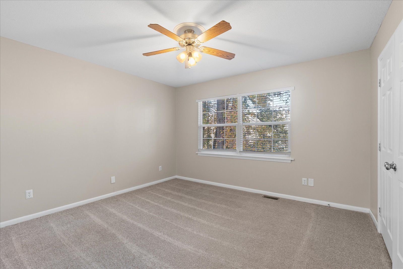 An empty room with neutral beige walls, grey wall-to-wall carpeting, a ceiling fan, and a window with white blinds.