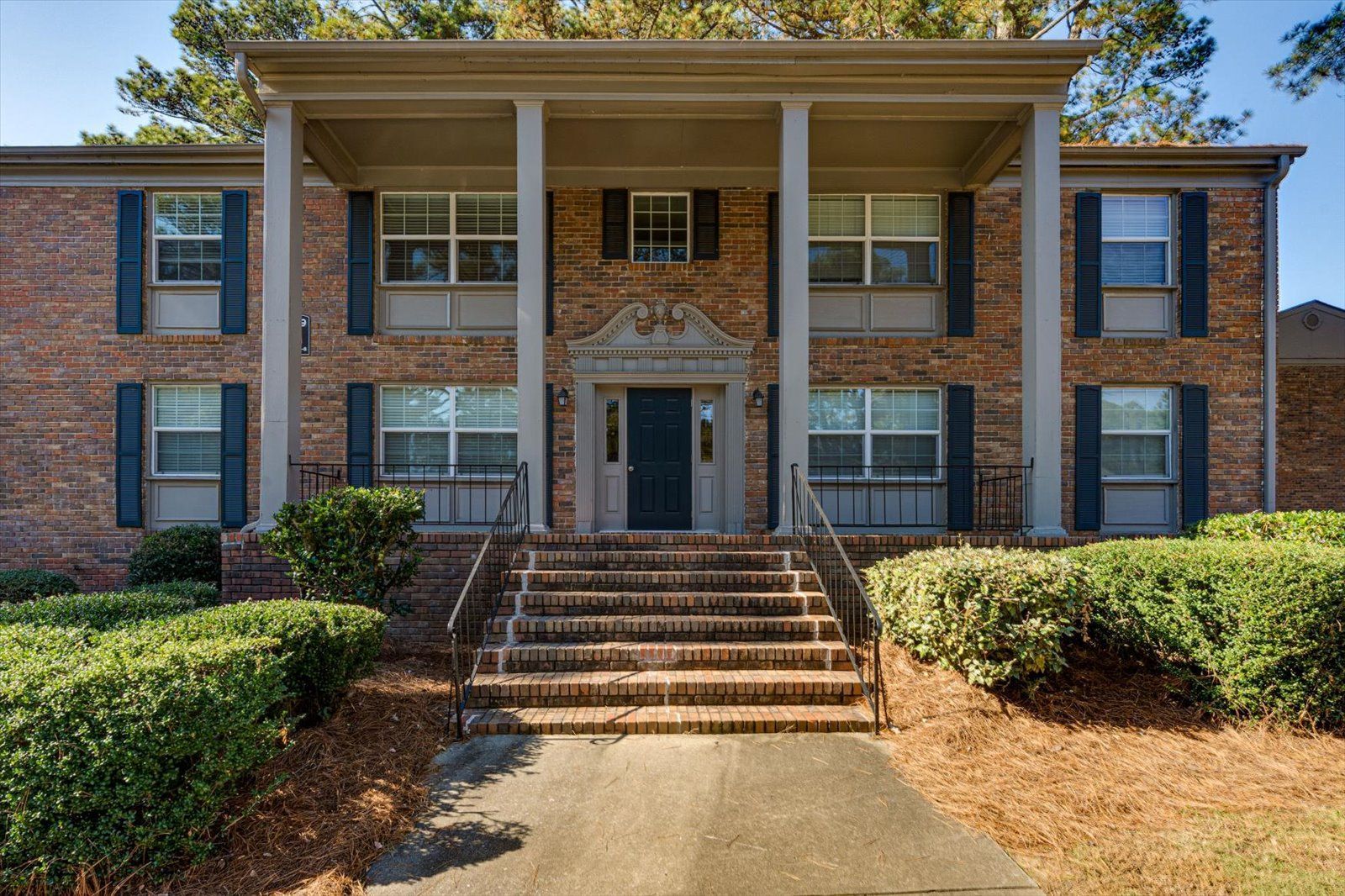 A two-story brick building with a columned front porch, a central dark door, and symmetrical windows with dark shutters.