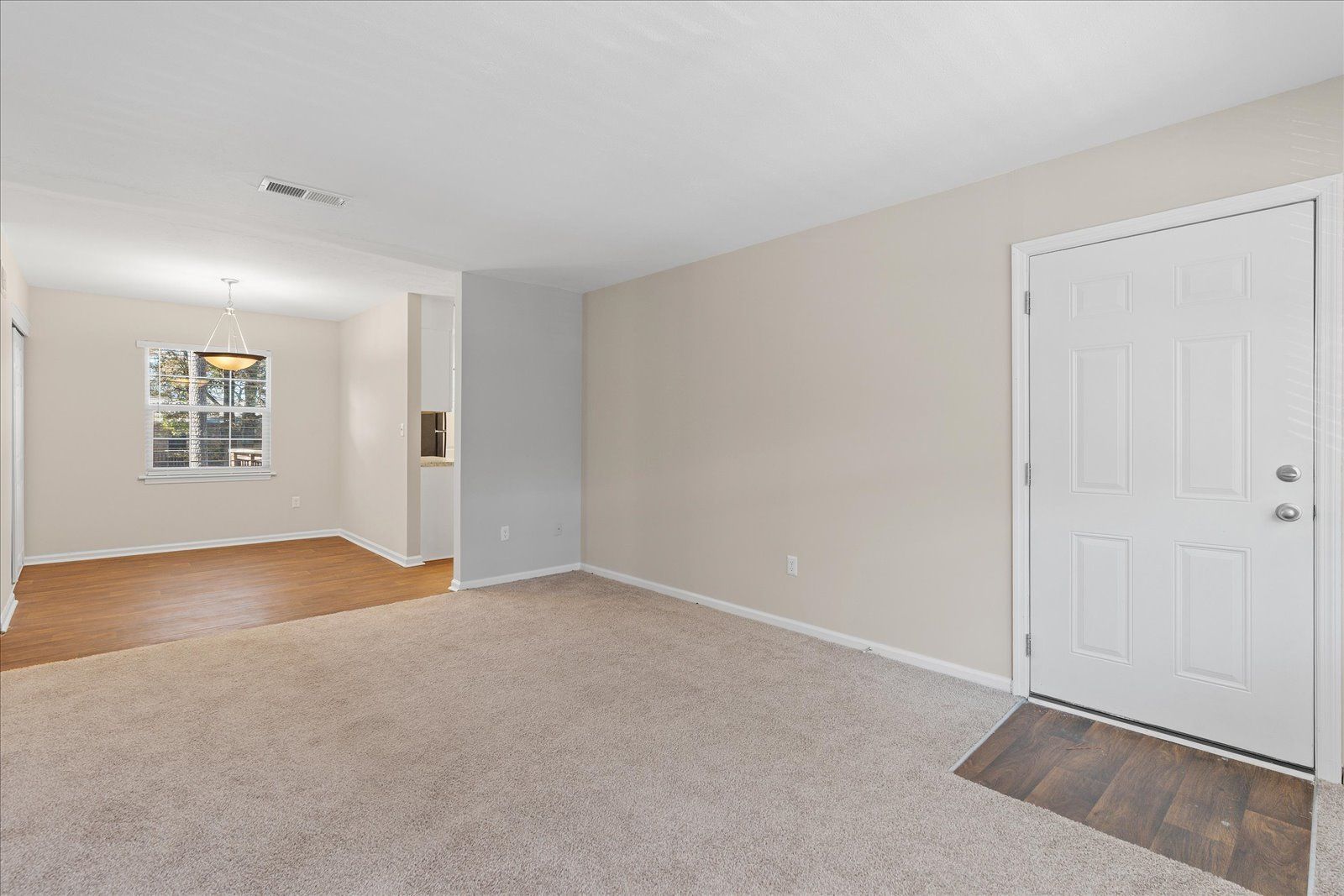 An empty room with beige carpet, light walls, a white front door, and a view into a dining area with a window.