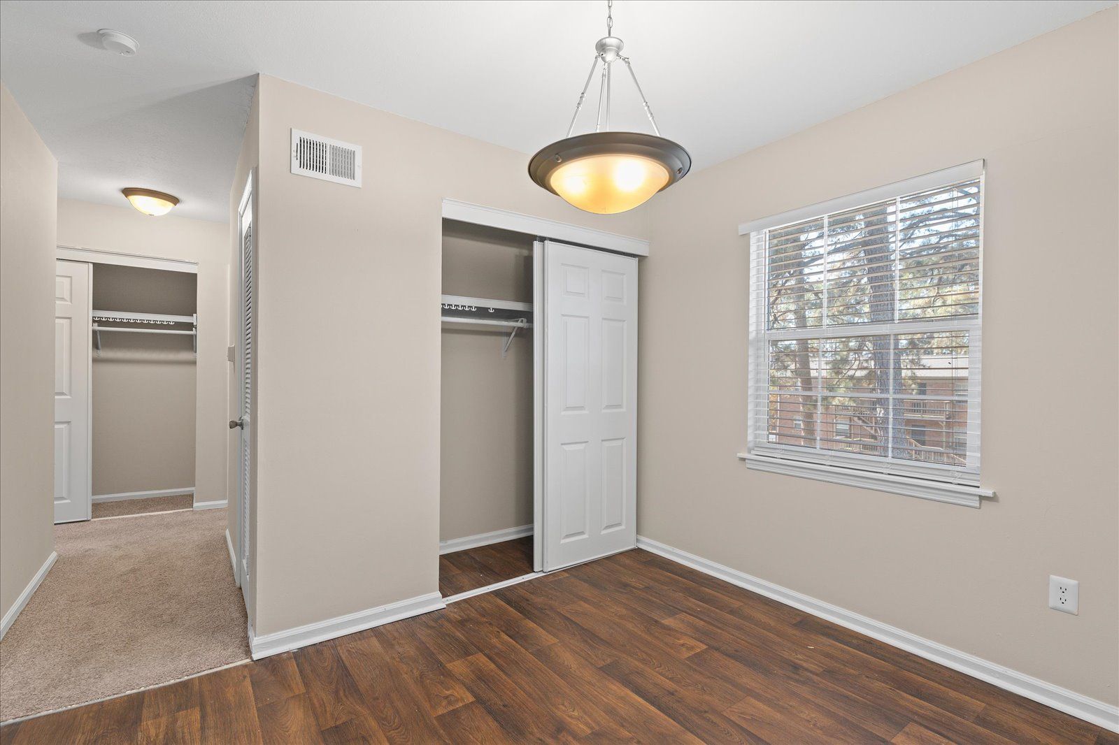 A light-walled room with wood-look flooring, a window, a pendant light, and two open closets.