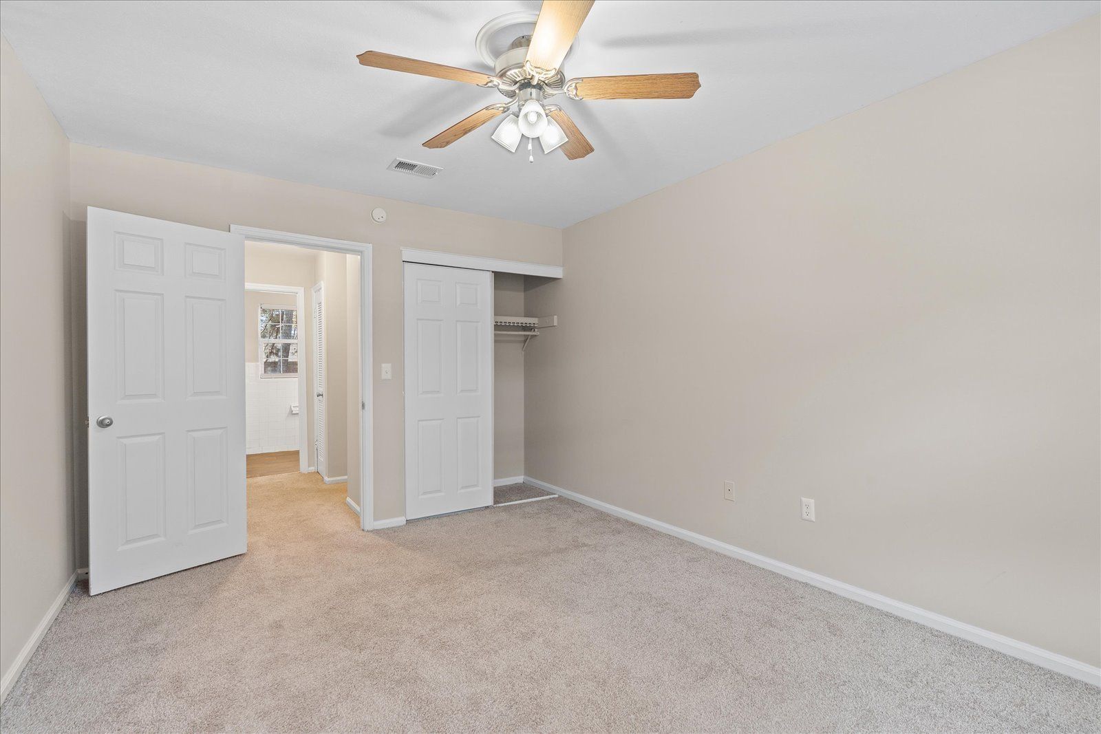 An empty bedroom with beige walls, light-colored carpet, a ceiling fan, and open white doors to a closet and hallway.