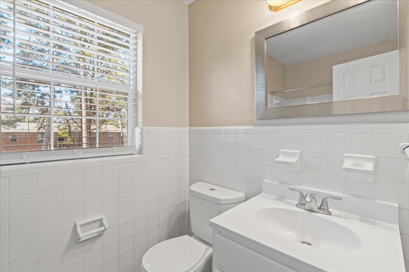 A bright bathroom featuring a white vanity with a silver faucet, a toilet, a rectangular mirror, and a window with blinds.