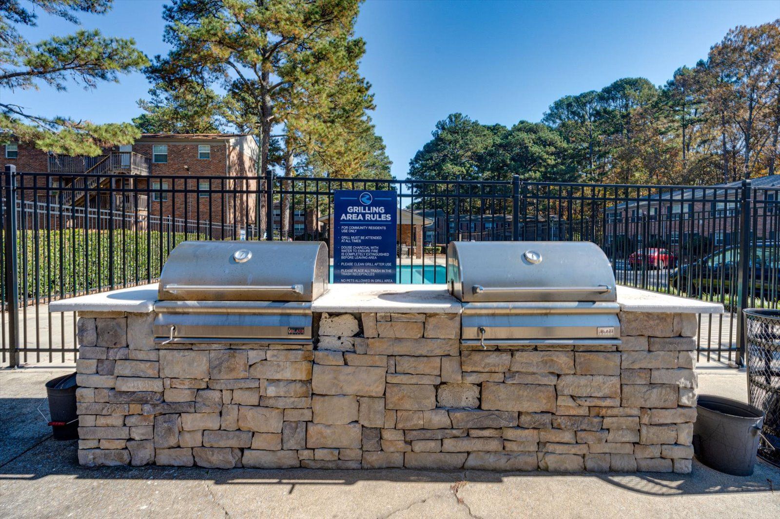 Two stainless steel outdoor grills set into a stone masonry counter near a black metal fence and swimming pool.