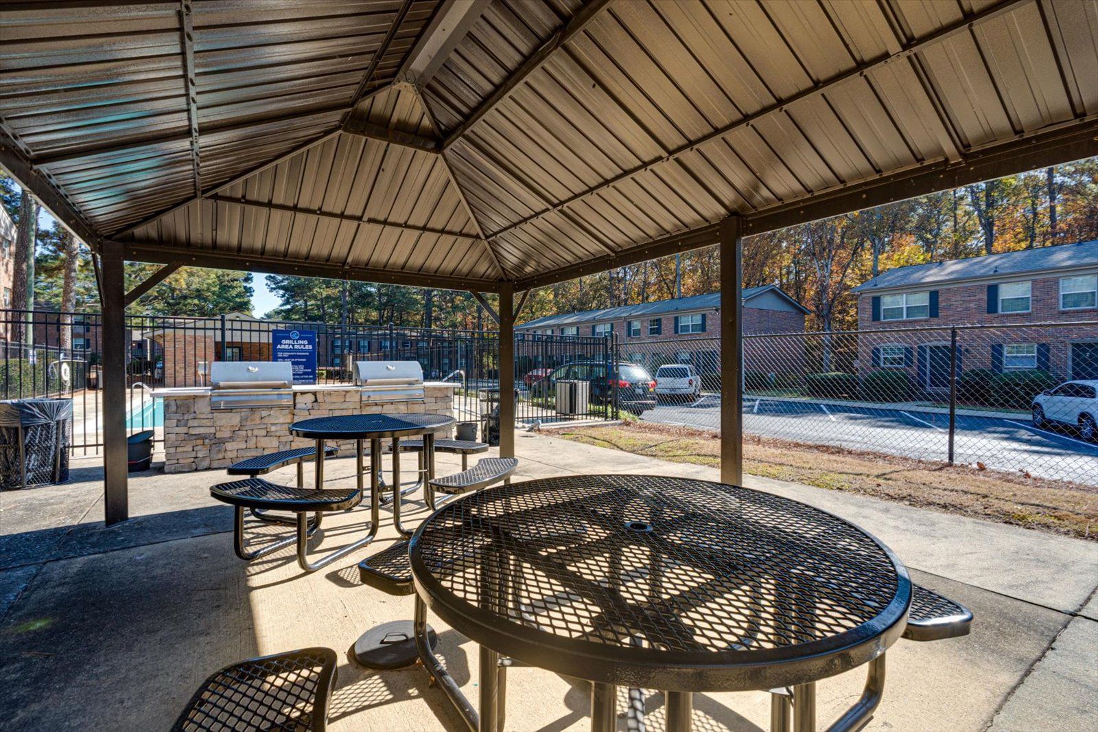 A metal gazebo with outdoor tables and a grilling area at an apartment complex parking lot.