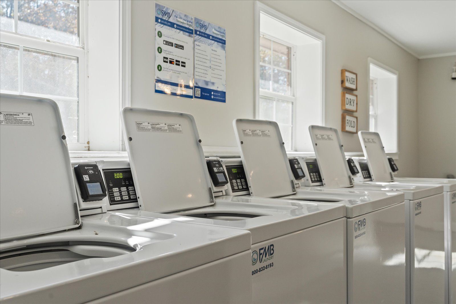 A row of white, top-loading washing machines with open lids in a brightly lit, clean laundry room.