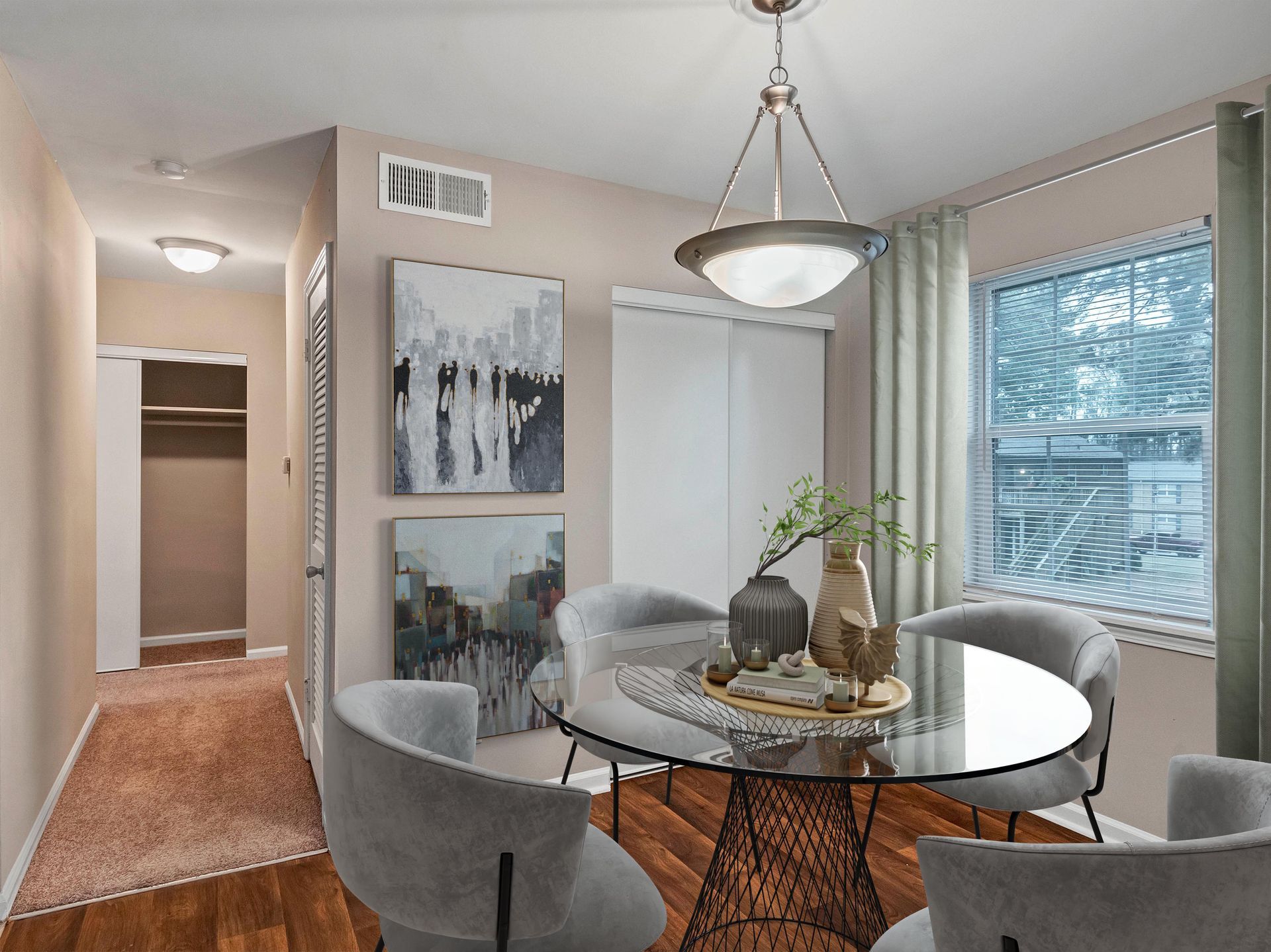 A round glass dining table with four grey upholstered chairs, pendant lighting, and wall art in a neutrally toned room.