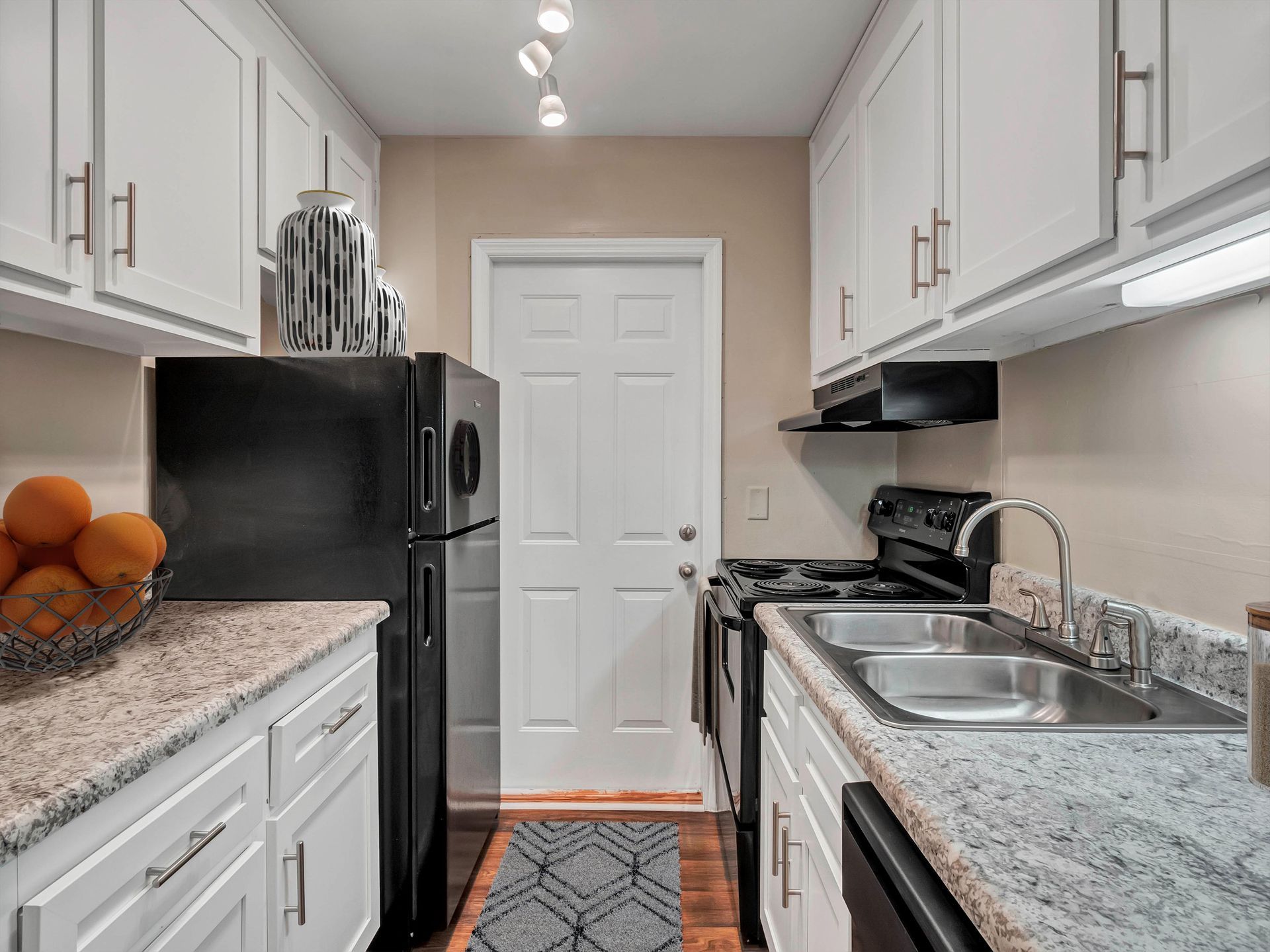 A narrow kitchen with white cabinets, speckled granite countertops, a black refrigerator, and a stainless steel sink.