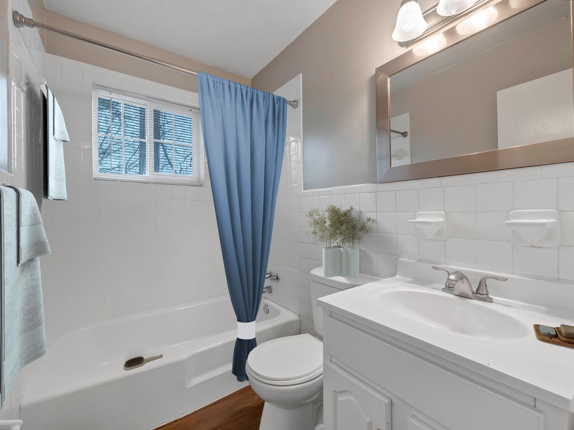 A bright bathroom featuring a white bathtub, a blue shower curtain, a pedestal sink, and a toilet against white tiled walls.