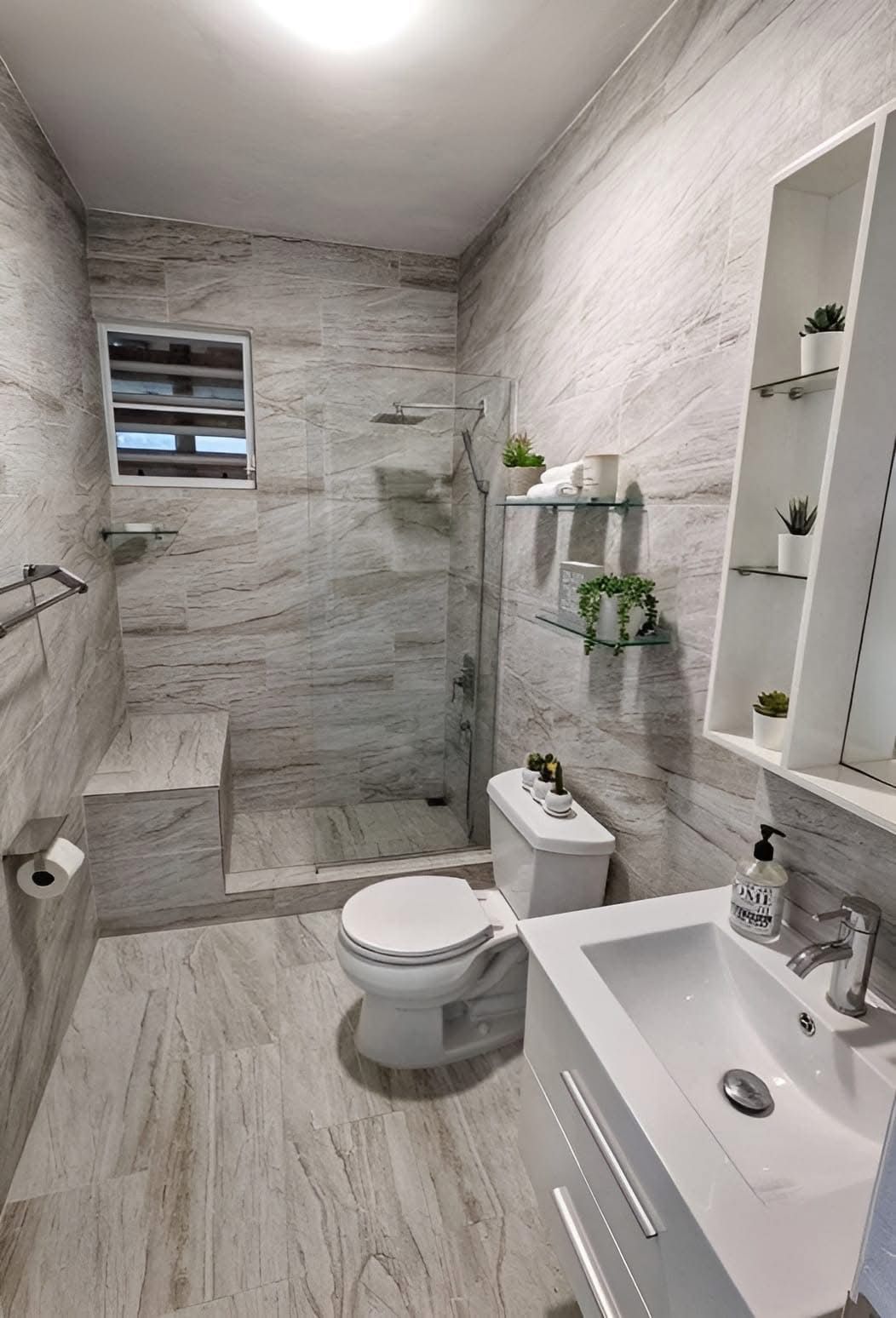 Modern bathroom with light grey stone-look tiles, a walk-in shower with a bench, a toilet, and a white vanity sink.