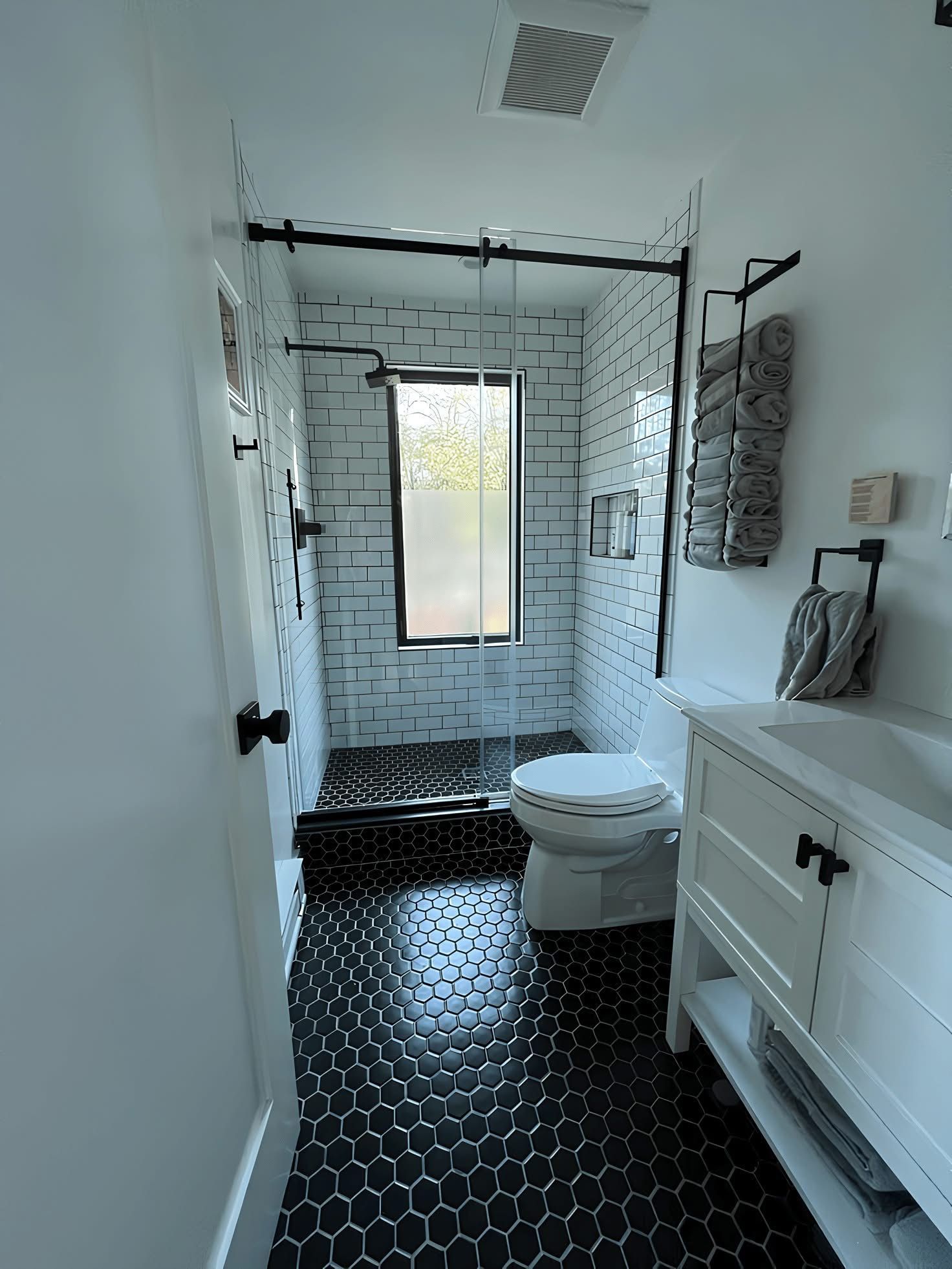 A small, modern bathroom featuring black hexagonal floor tiles, white brick-patterned shower walls, and a white vanity.