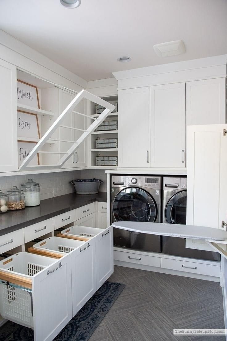 A bright, organized laundry room with white cabinets, pull-out laundry baskets, a fold-down drying rack, and ironing board.