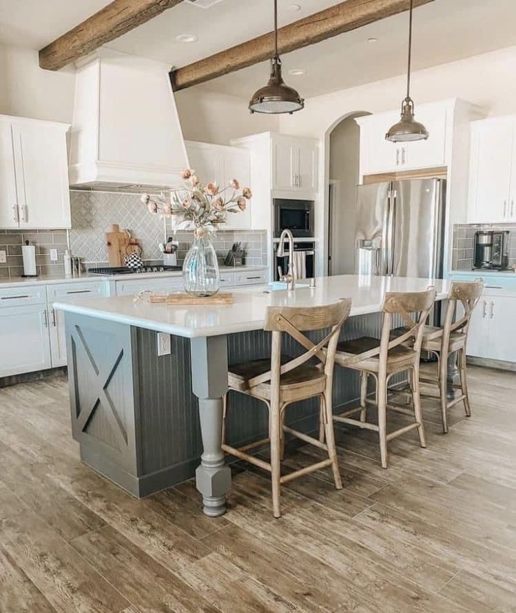 A bright, modern farmhouse kitchen featuring a grey island with cross-brace details, white cabinets, and wooden beams.