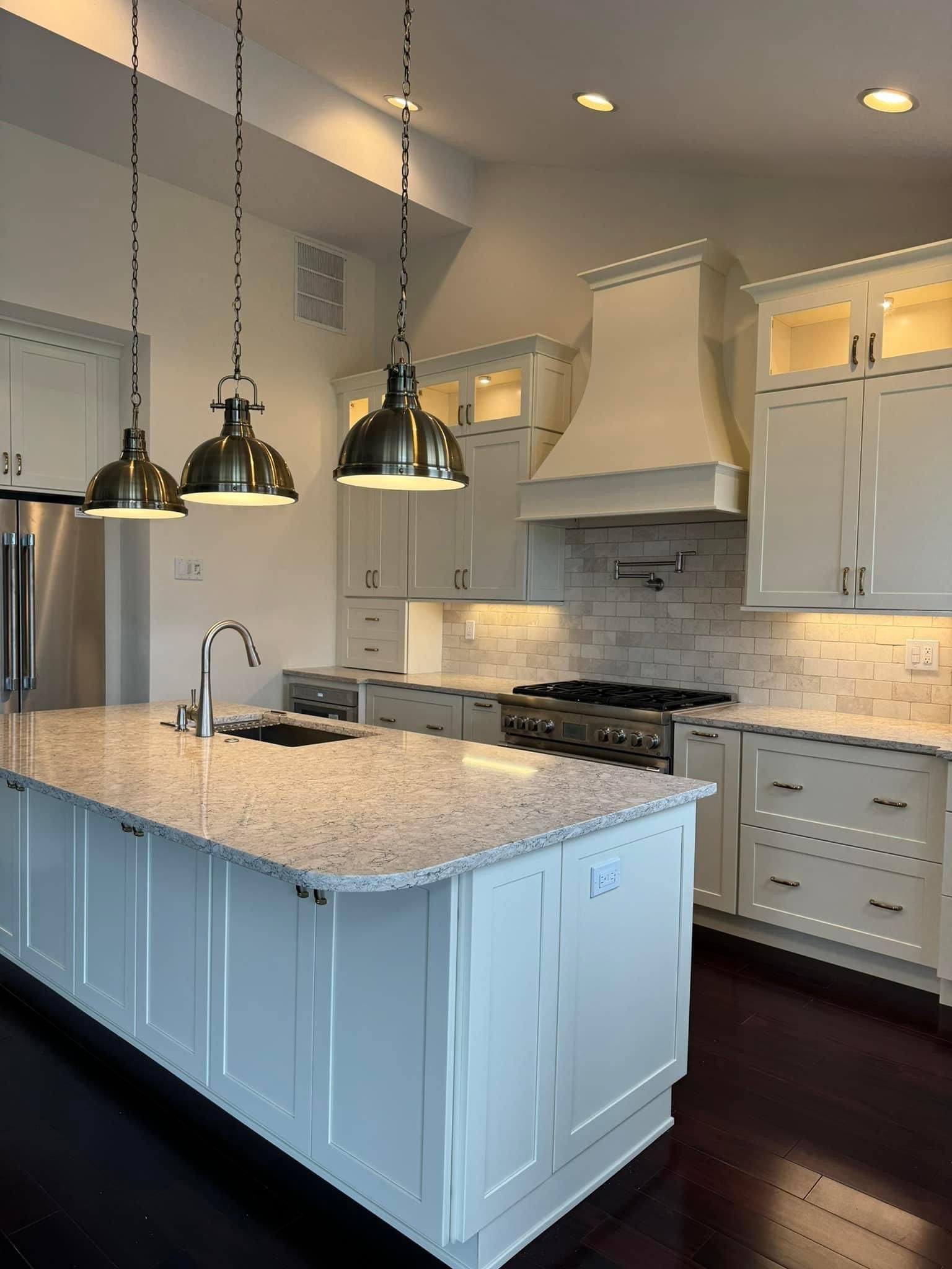 A bright, modern kitchen featuring white cabinets, a large granite island with pendant lights, and dark wood floors.