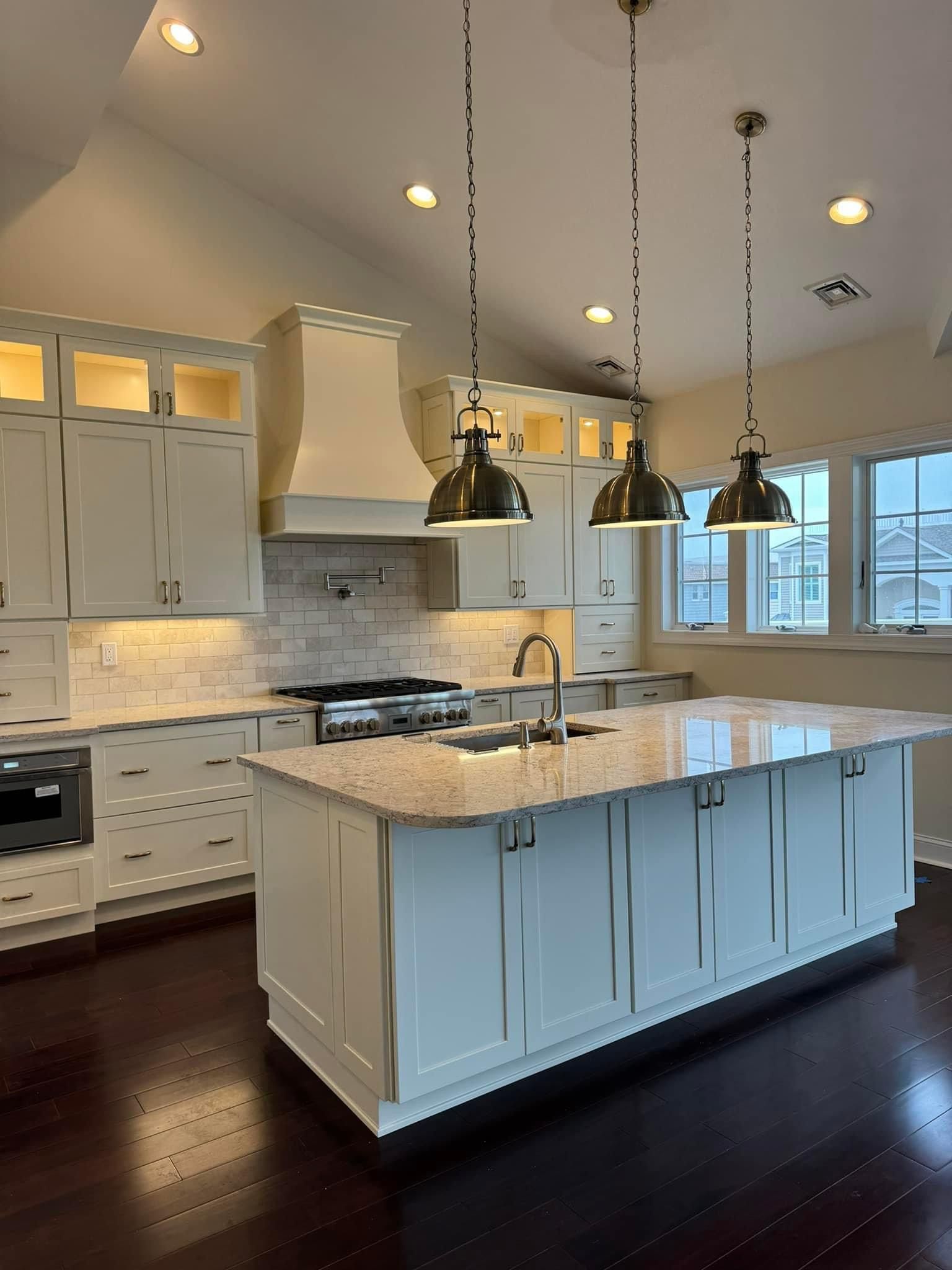 A bright kitchen with white cabinets, a large granite-topped island, dark hardwood floors, and three metal pendant lights.