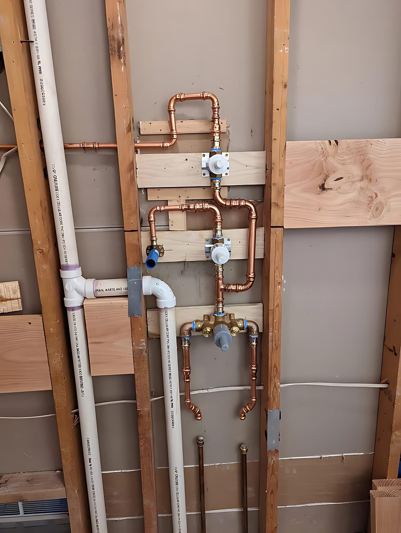 Copper plumbing pipes and valves installed on wooden wall studs next to white PVC drain pipes in an unfinished wall.