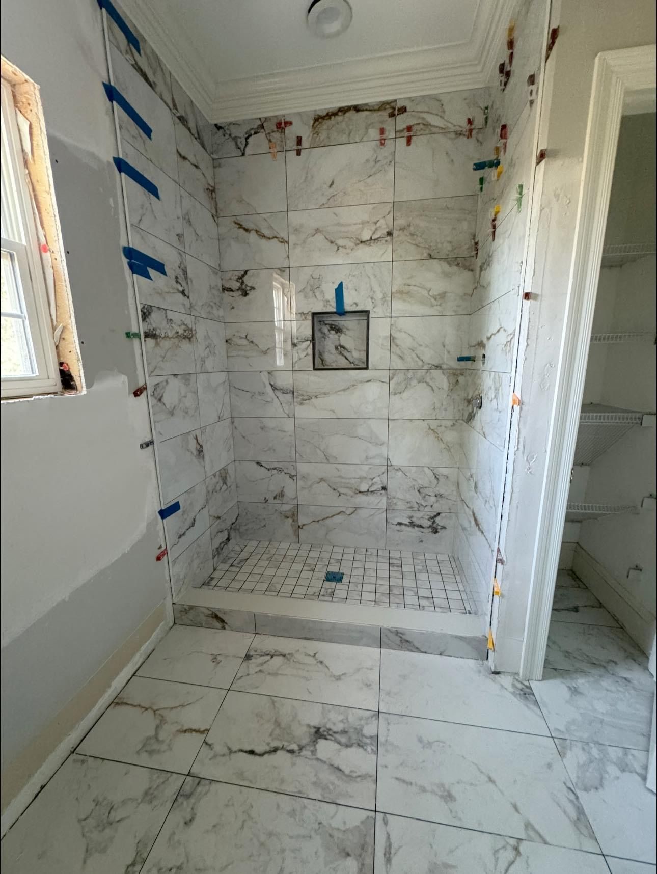 A bathroom shower under renovation with white marble-patterned tiles on the walls and floor and an inset storage niche.