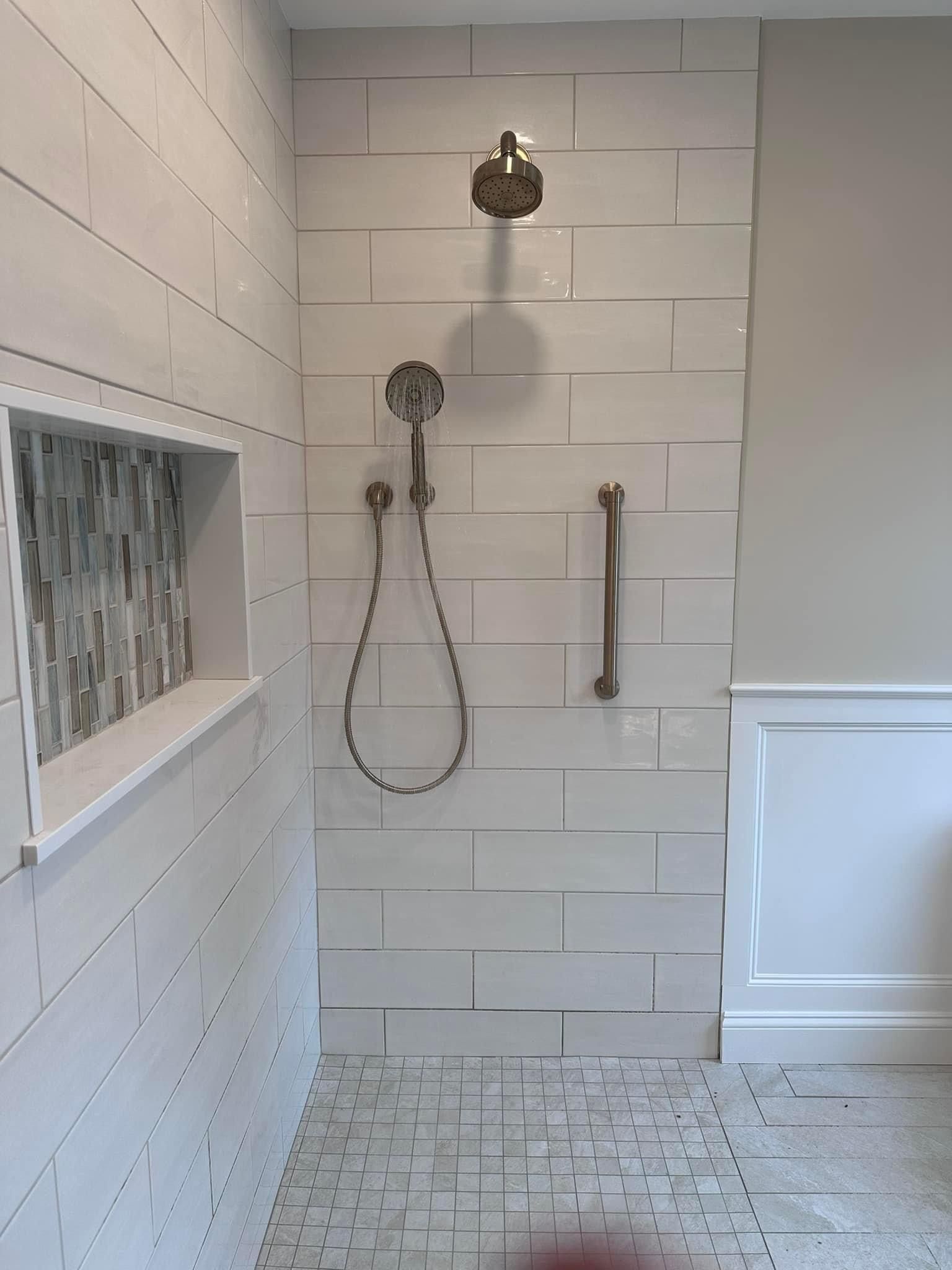 A walk-in shower with a rainfall showerhead, a handheld sprayer, a grab bar, and a recessed tiled niche on a white wall.