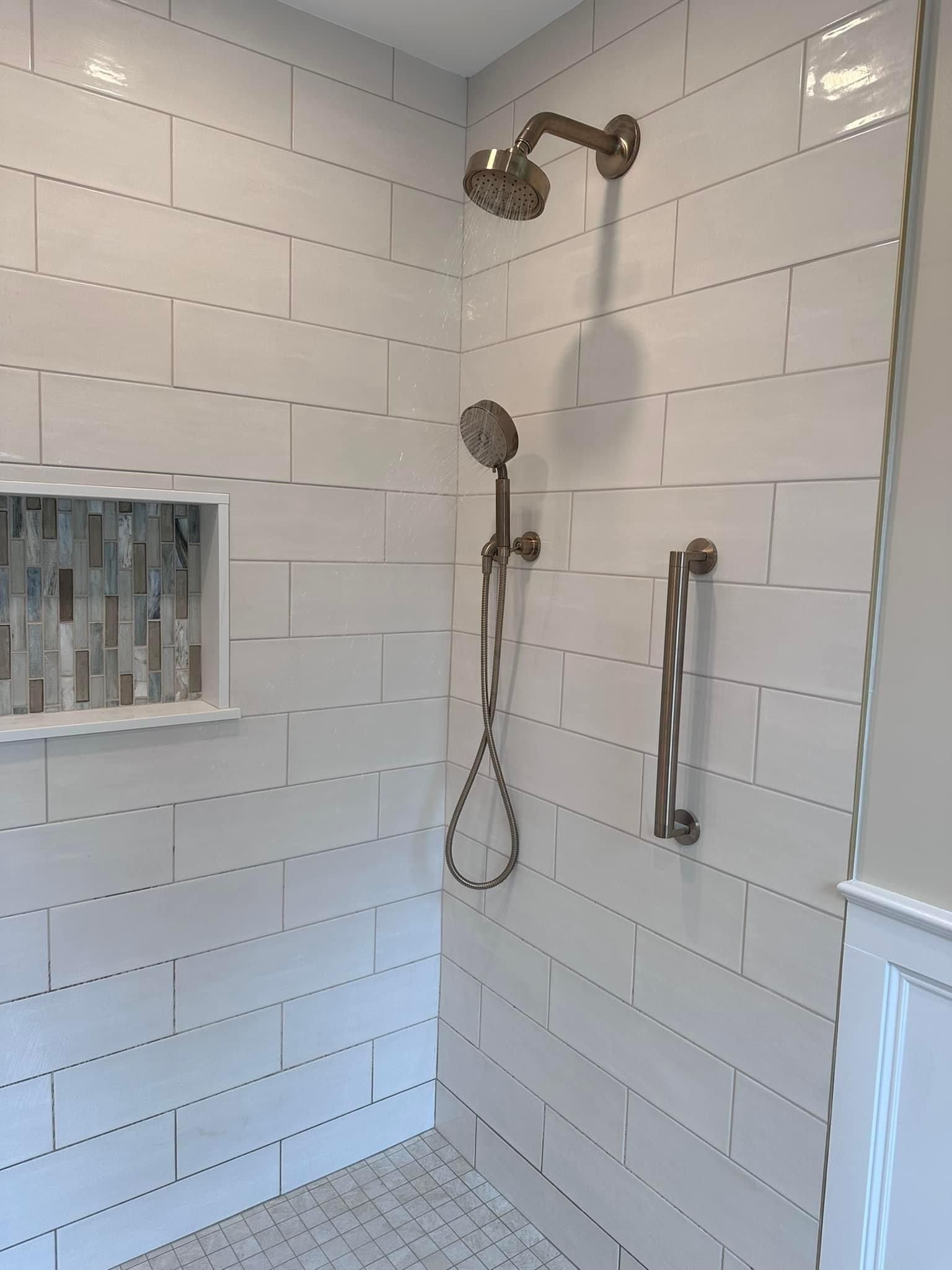 A shower interior with white rectangular tiles, a tiled recessed niche, a bronze shower head, a handheld sprayer, and bar.