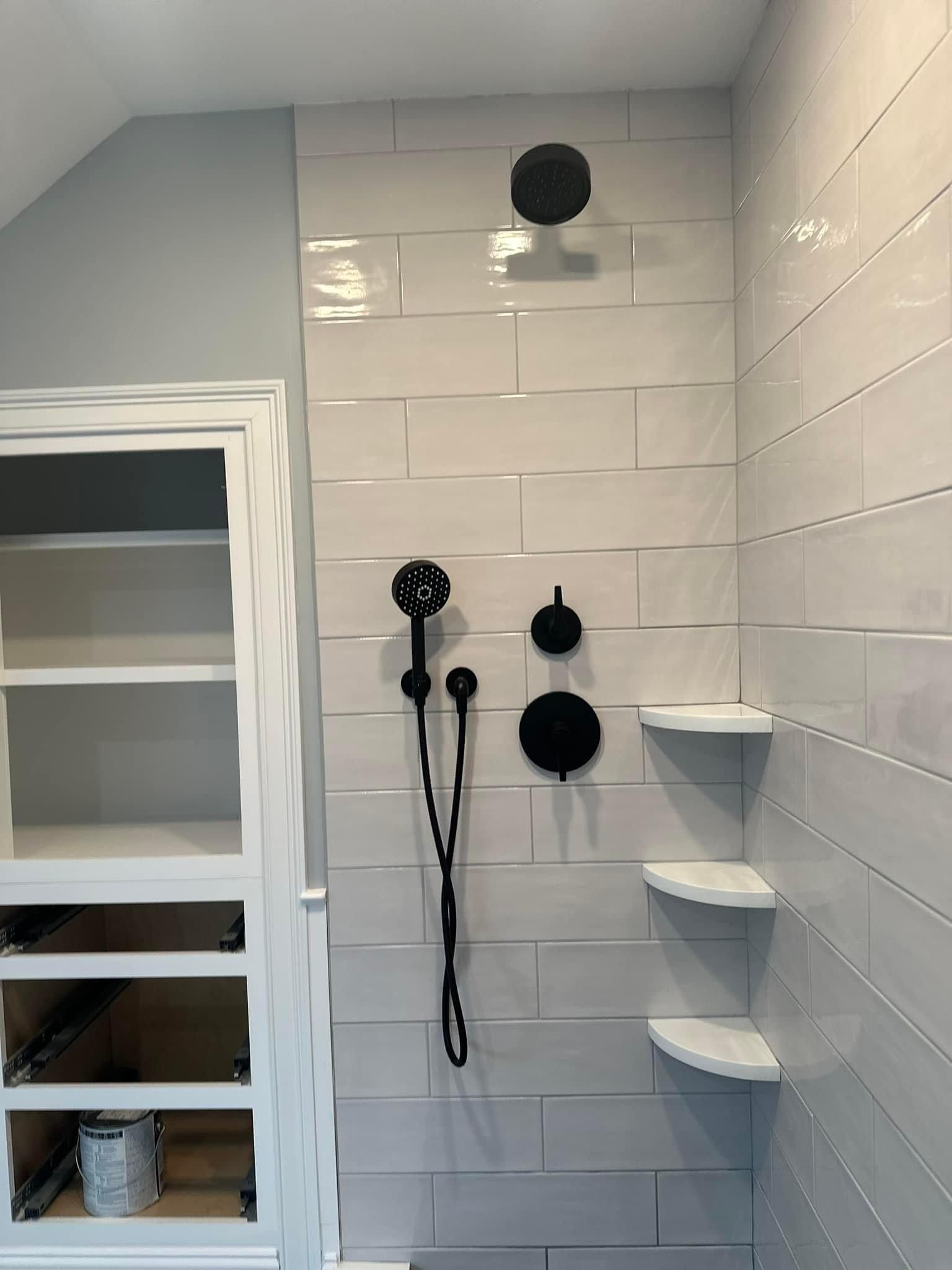 A modern shower featuring large light-colored tiles, black fixtures, a handheld sprayer, and three built-in corner shelves.