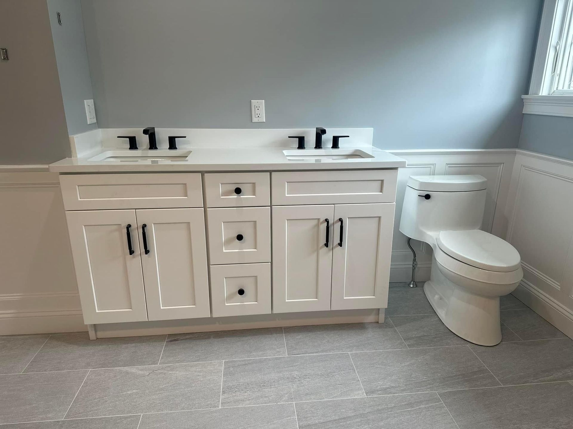 A modern bathroom with a double vanity, white cabinets, black hardware, and a white toilet against light gray walls.