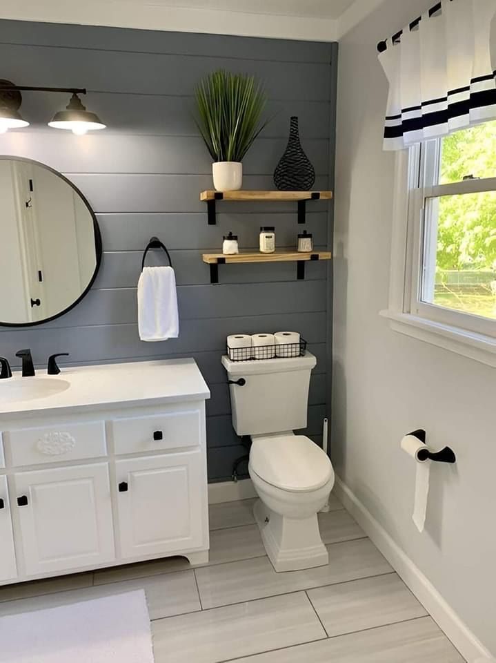 A modern bathroom with a blue shiplap accent wall, white vanity, round mirror, and two floating wooden shelves above toilet.