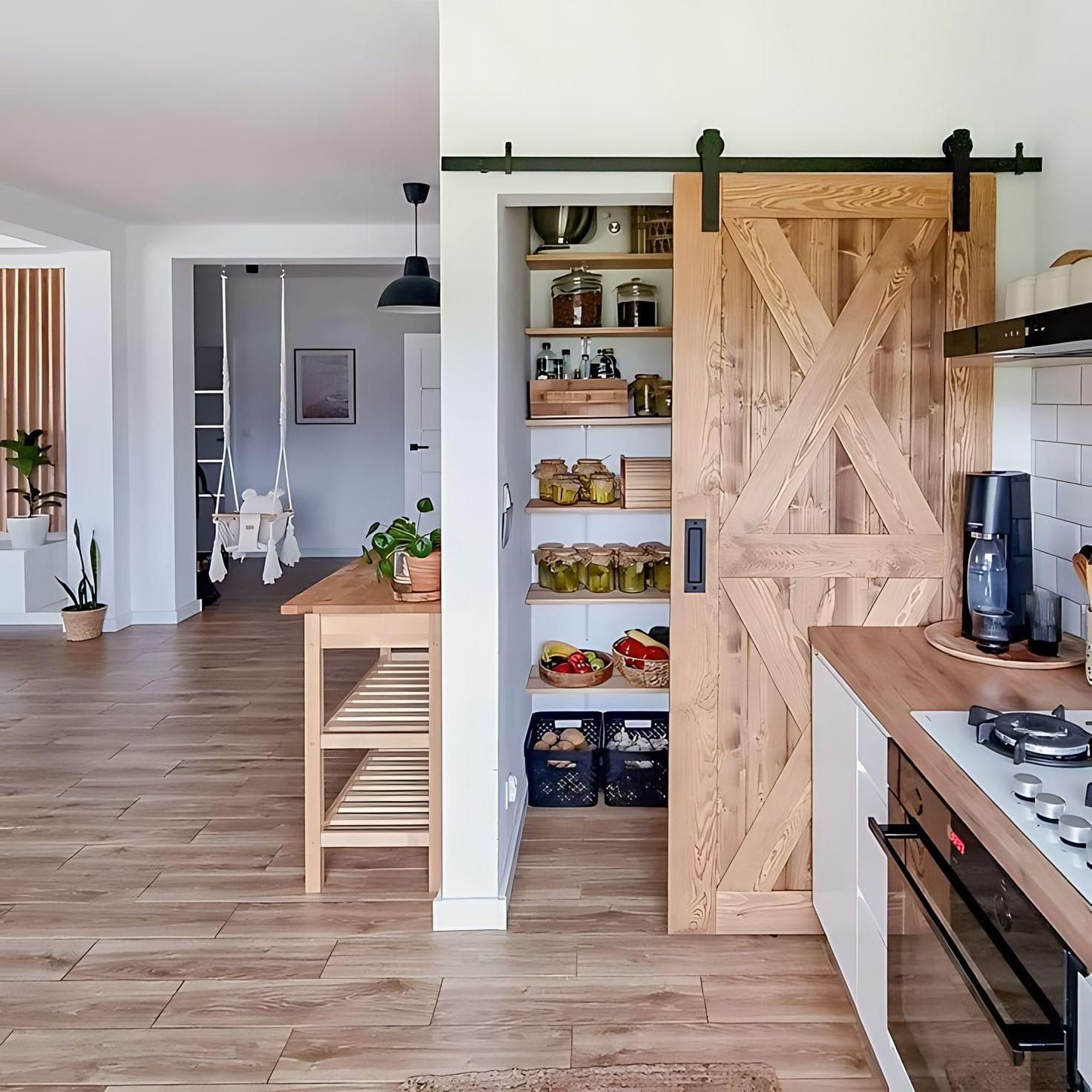 A wooden barn-style door slides open to reveal a kitchen pantry with shelves of jars and baskets, next to a modern stove.