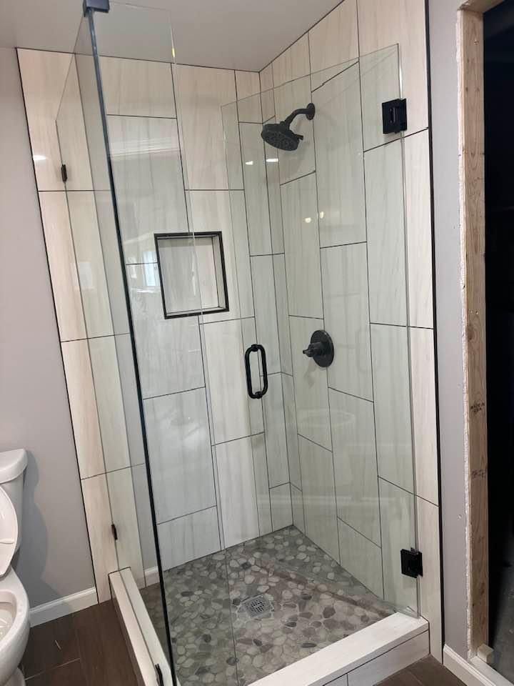 A glass-enclosed shower with off-white subway tile walls, a pebble-stone floor, and black fixtures.