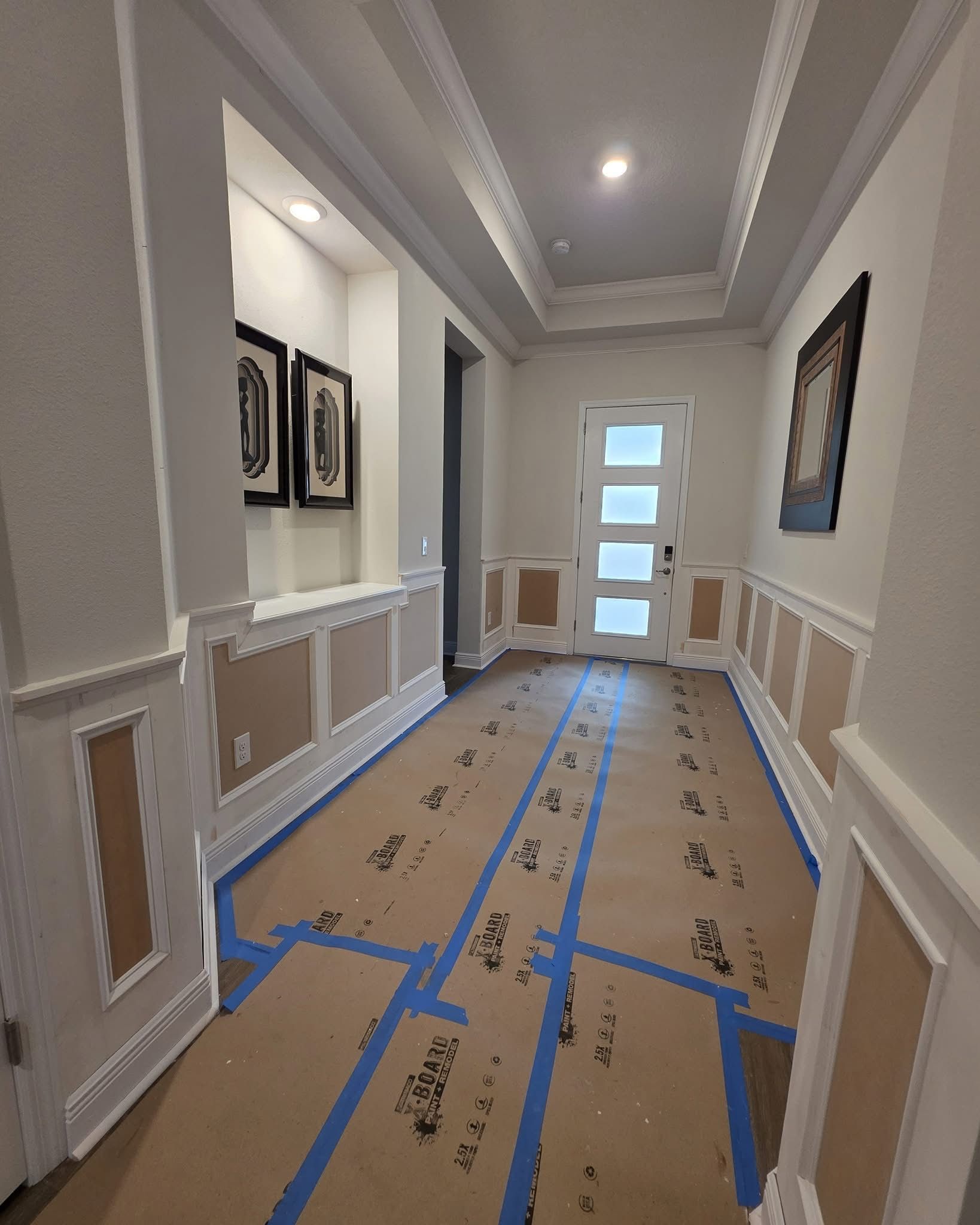 A hallway under renovation with white wainscoting and molding, beige paper covering the floor, and framed wall art.