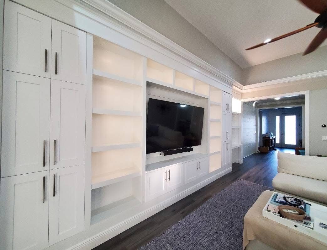A white built-in wall unit with shelving and a mounted TV in a living room, viewed near a sofa and doorway.