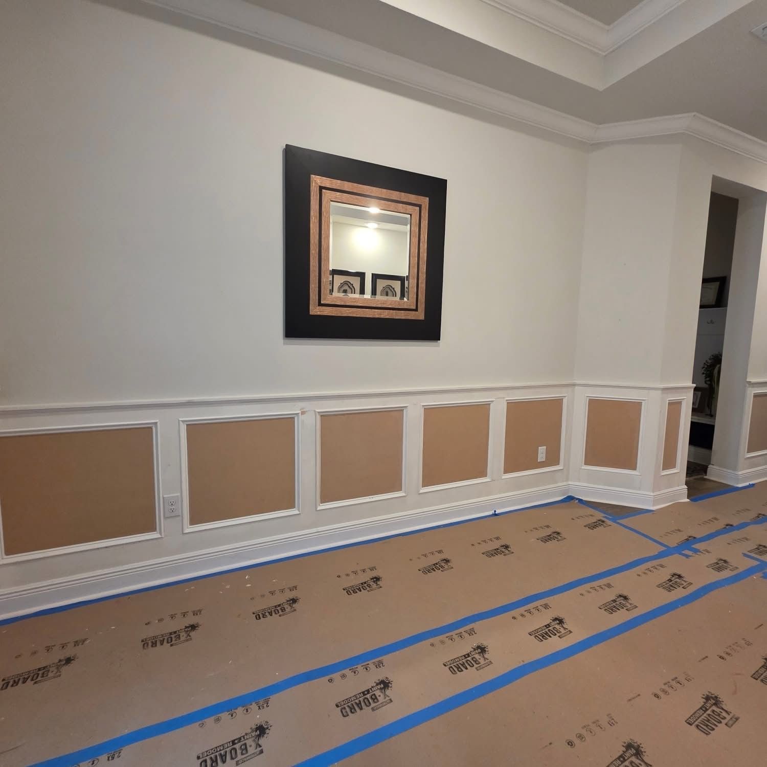 A hallway with white wainscoting and a mirror on the wall, with brown protective paper covering the floor.