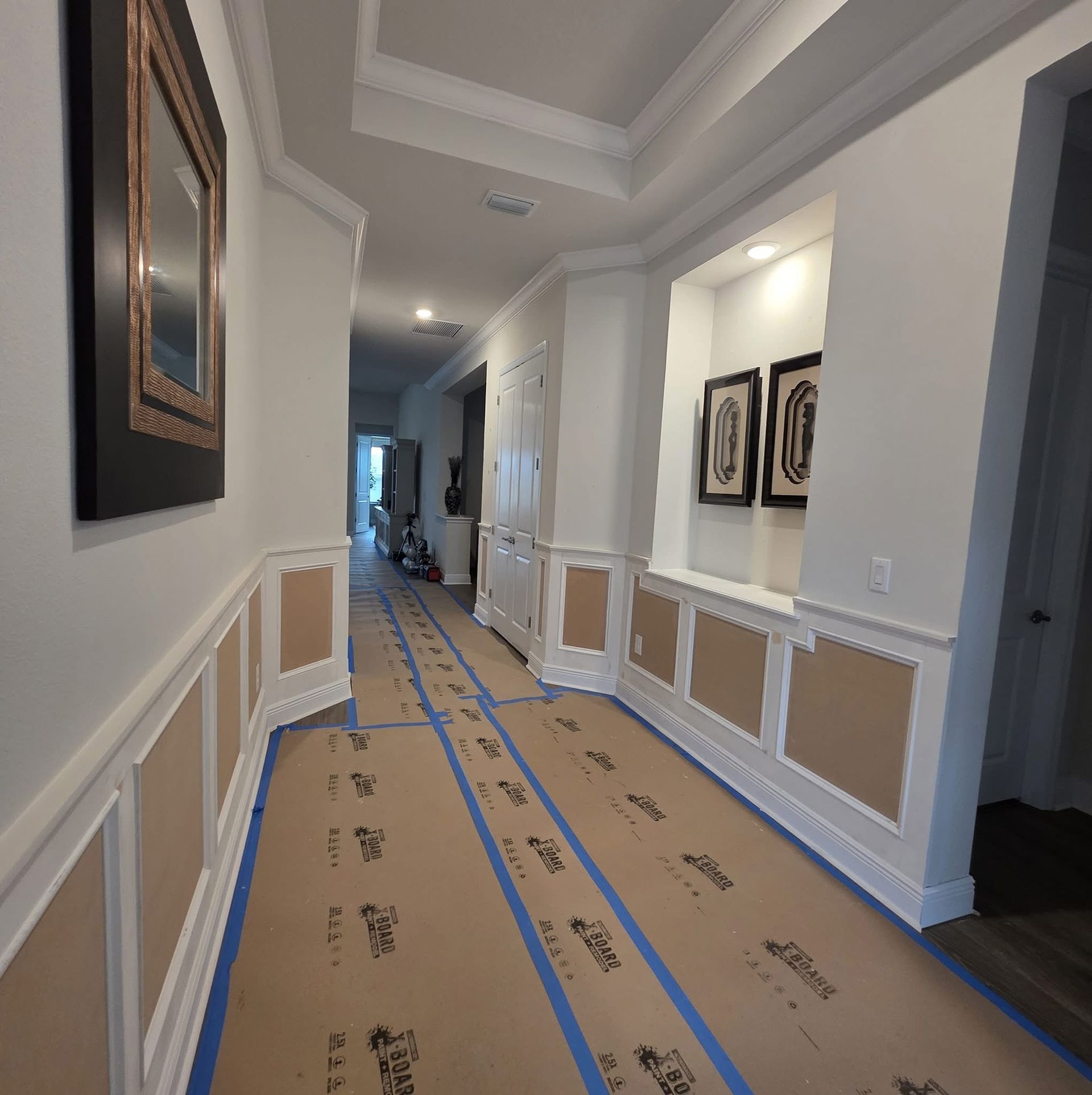 A hallway with white walls and wood-paneled wainscoting, featuring protective paper floor covering secured by blue tape.