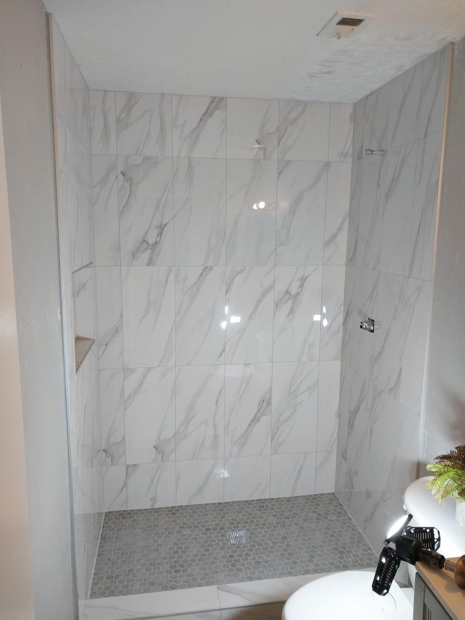 A modern walk-in shower with white marble-patterned wall tiles and a small grey pebble floor, viewed from a bathroom.