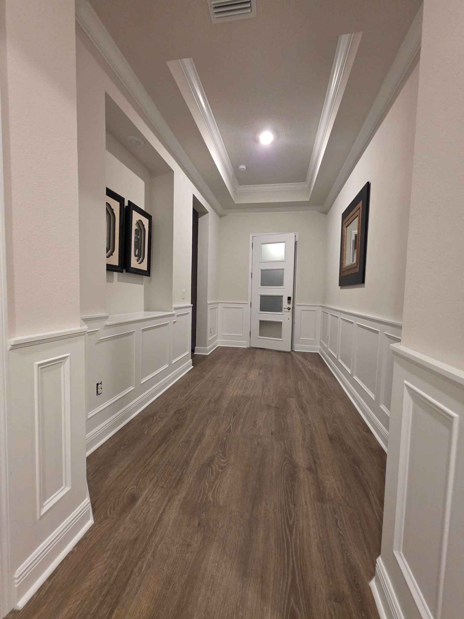 A brightly lit hallway with white wainscoting, wood-look flooring, wall art, and a white door at the far end.