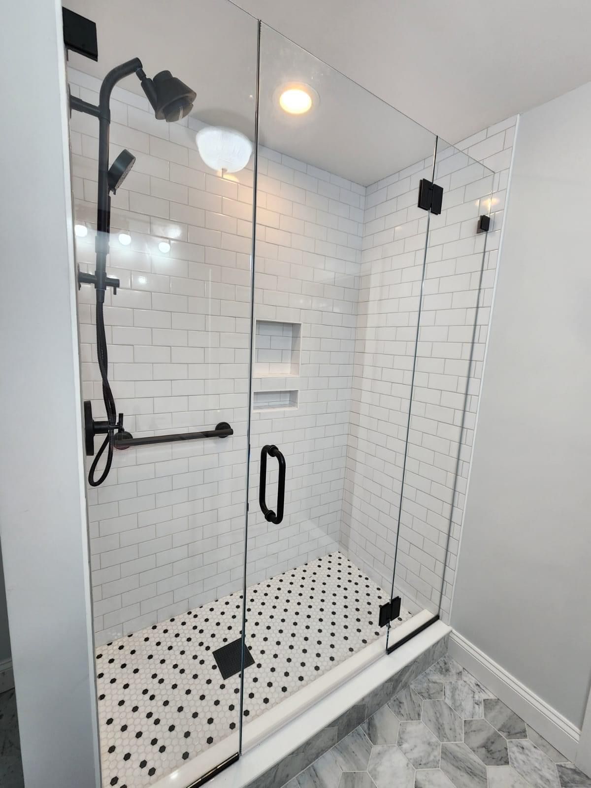 A glass-enclosed shower with white subway tile walls, a matte black shower head, and a black-and-white patterned floor.