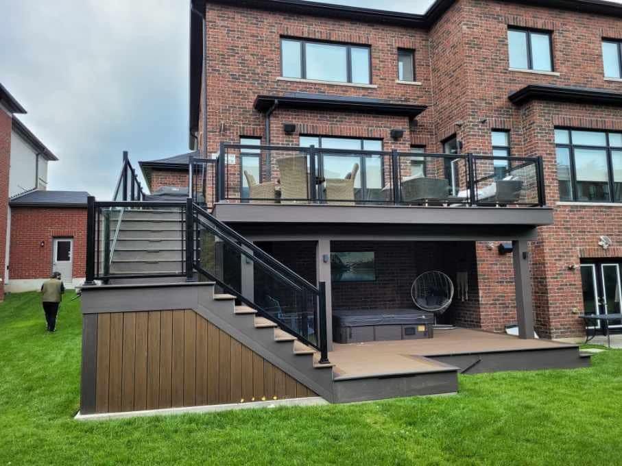 A two-story brick house with a raised, dark-toned composite deck, glass railings, stairs, and a covered patio area.