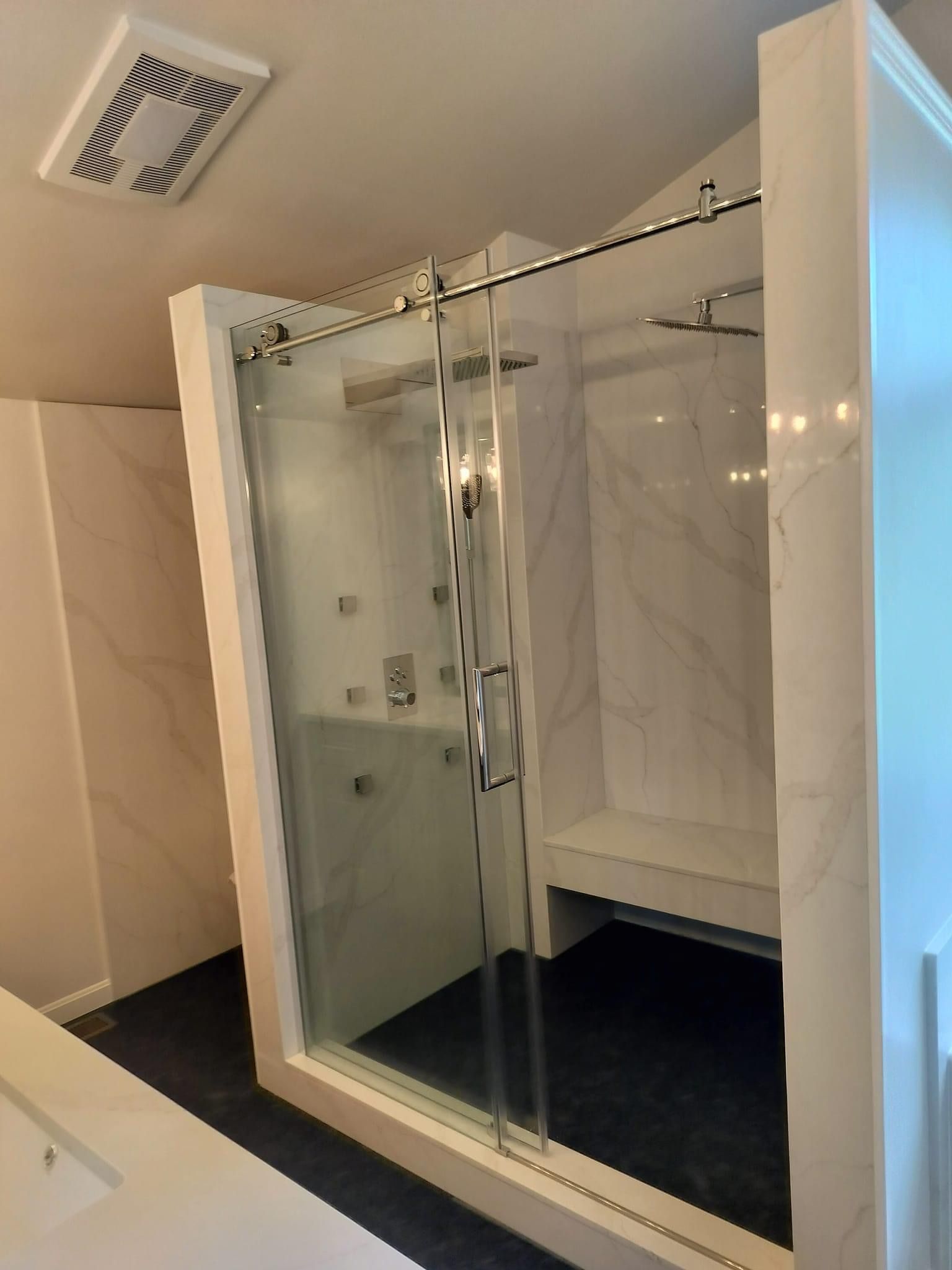 A walk-in shower with white marble-patterned walls, a dark floor, a built-in bench, and a glass sliding door.