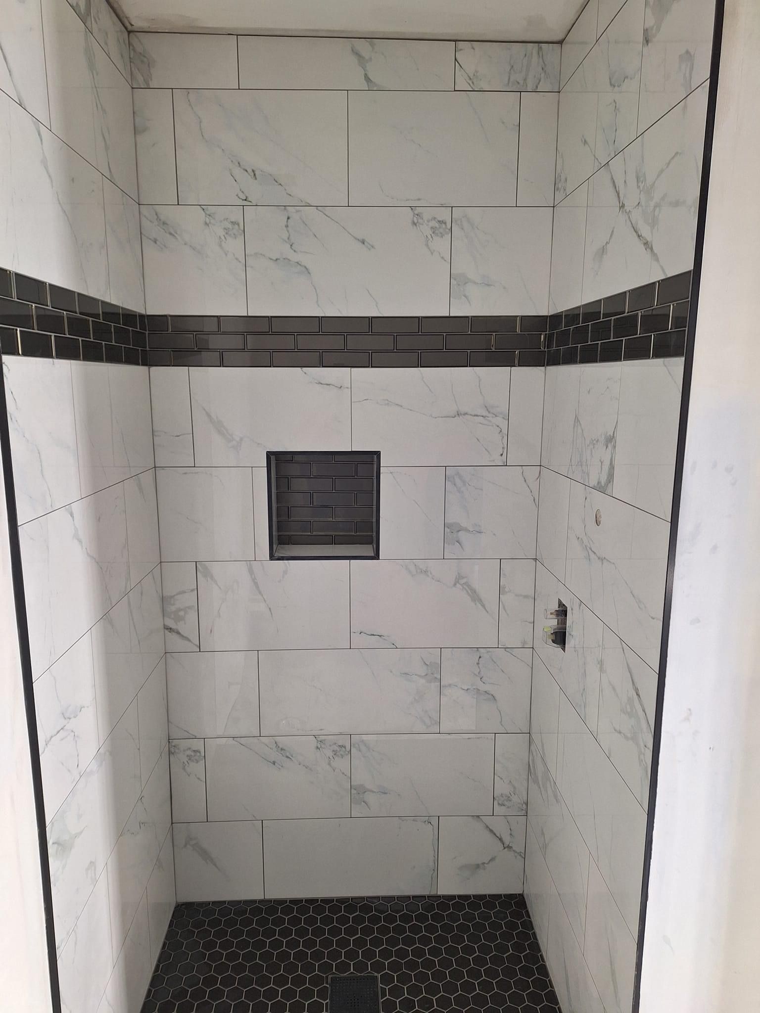 A shower enclosure with white marble-look tiles, a dark horizontal accent stripe, a matching dark niche, and pebble floor.