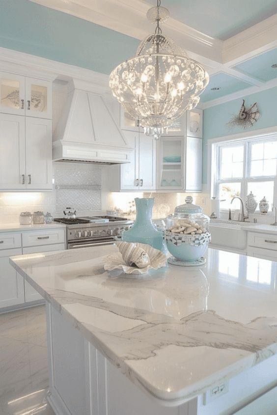 A bright, white coastal-style kitchen with a marble-topped island, light blue ceiling, and ornate crystal chandelier.
